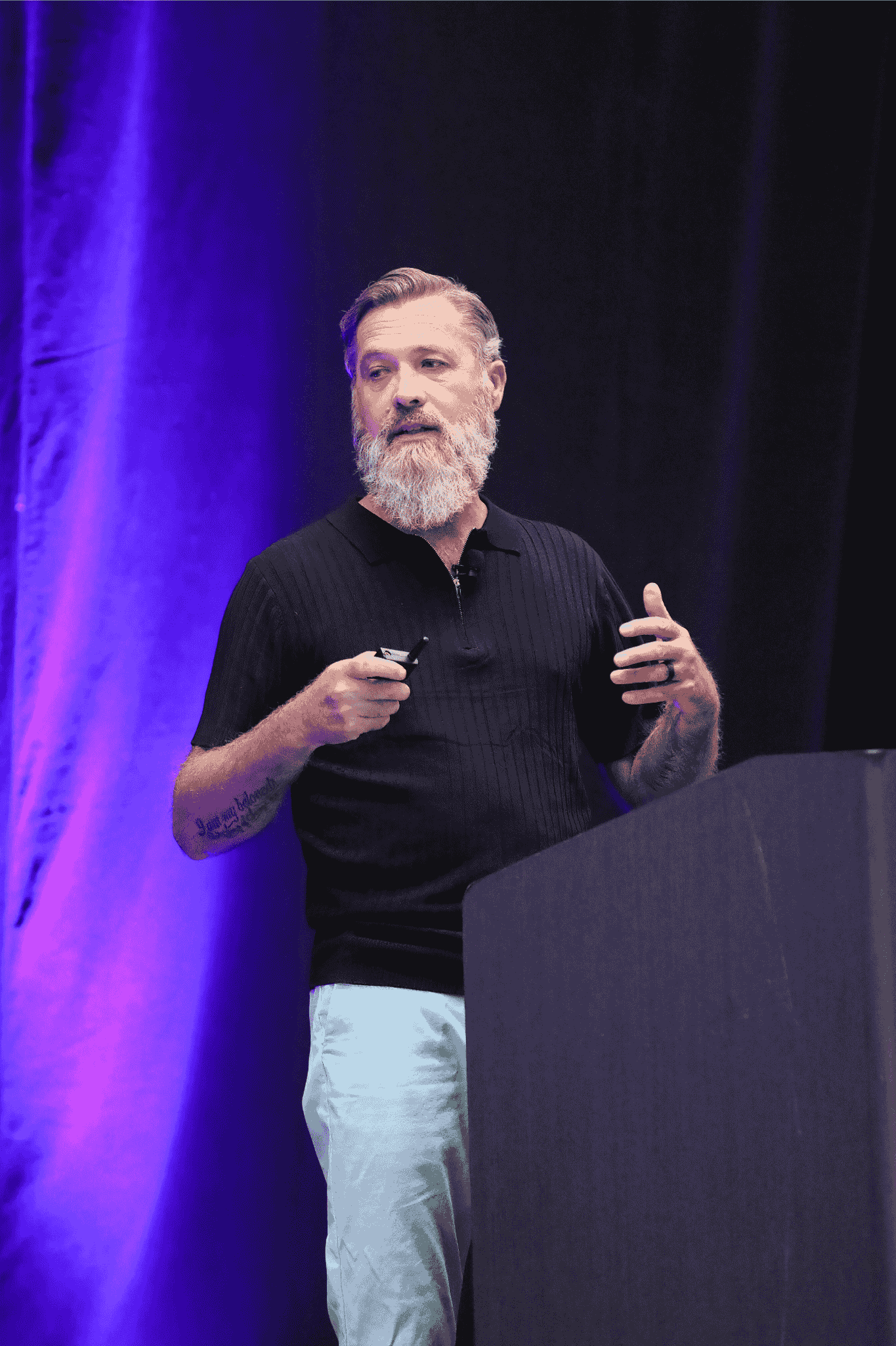 ダラスで開催されたIQGeo Americas Meetup 2025のステージで、紫色のステージ照明の下、満員の聴衆を前にプレゼンテーションを行うClearwave Fiber社のアラン・ダージー氏。