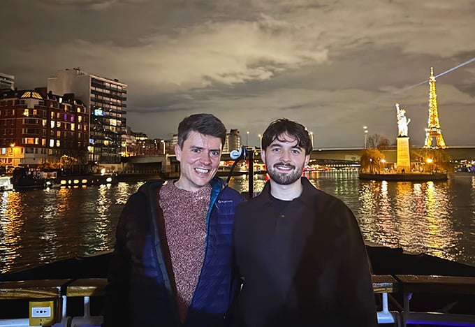 夜の川辺の遊歩道に二人が立っている。背後には明かりの灯った街のビル群が広がる。対岸の小さな島には、曇り空の下で明かりを灯した塔状の建造物と像が見える。