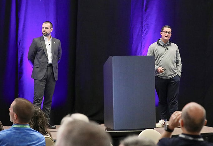 紫色の照明が当たる背景の前で、二人の司会者がステージ上に立っている。一人が話し、もう一人が聞いている。